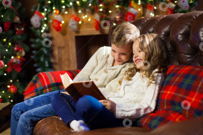 Скачать Дети читают интересную книгу, сидя на кровати на фоне украшенной рождественской елки фотосток Ozero