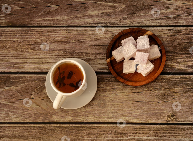 Скачать Чашка горячего травяного чая на блюдце и круглая деревянная тарелка с кучей восточных сладостей в сахарной пудре на деревянном столе. фотосток Ozero