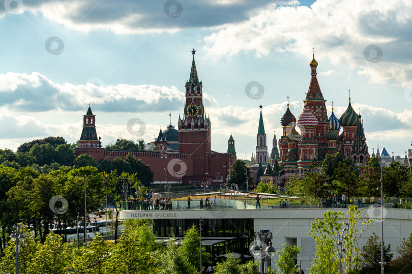 Скачать вид на Спасскую башню Московского Кремля и собор Василия Блаженного из парка Зарядье на фоне голубого неба. Люди на смотровой площадке здания фотографируются. фотосток Ozero