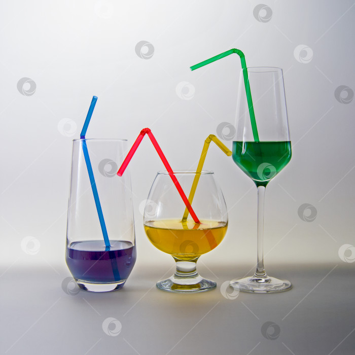 Скачать Три бокала с цветными напитками стоят в ряд, в них вставлены разноцветные трубочки для коктейлей, на светло-сером фоне. фотосток Ozero