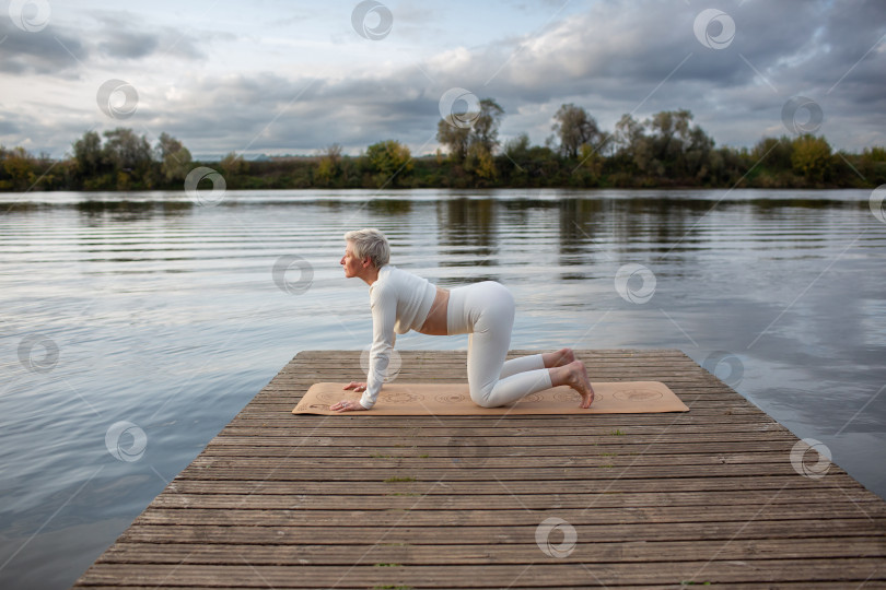 Скачать Взрослая женщина в белом костюме выполняет асаны йоги на пирсе у реки. Маджариасана. Поза кошки, наклонившись фотосток Ozero