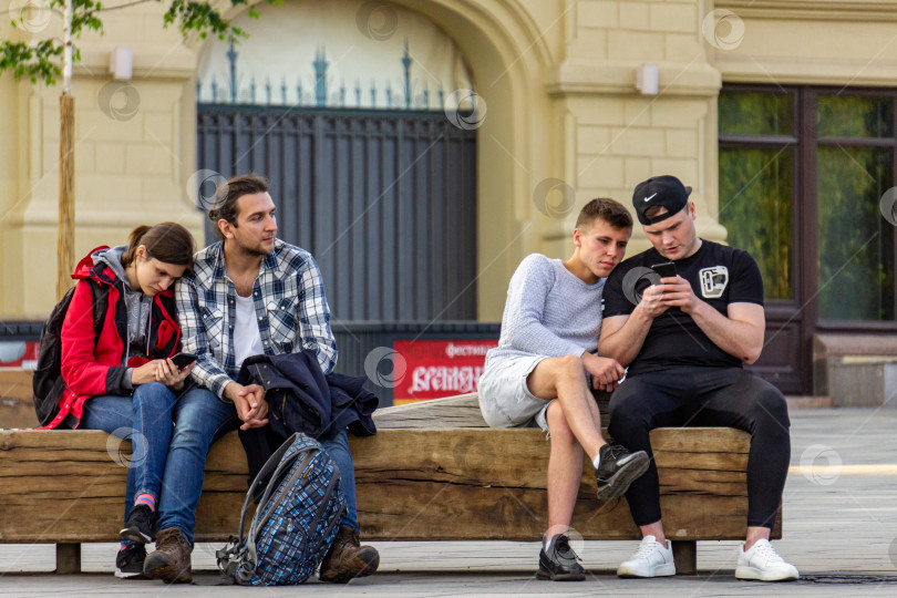 Скачать Молодые люди сидят на скамейке в парке Зарядье и отдыхают. Девушка печатает на телефоне.  Двое молодых людей внимательно смотрят на экран смартфона. фотосток Ozero