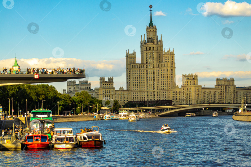 Скачать Прогулочные катера стоят у причала на фоне наплавного моста и сталинской высотки.Люди прогуливаются по наплавному мосту. фотосток Ozero