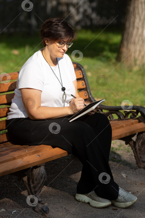 Скачать Красивая женщина средних лет сидит на скамейке, записывая идеи в блокнот фотосток Ozero