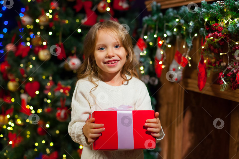 Скачать Маленькая девочка сидит у елки с рождественским подарком в руках фотосток Ozero