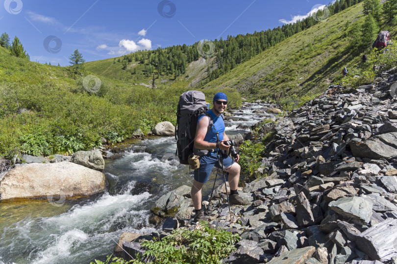 Скачать Счастливый молодой турист с рюкзаком и фотоаппаратом стоит на берегу горной реки. фотосток Ozero