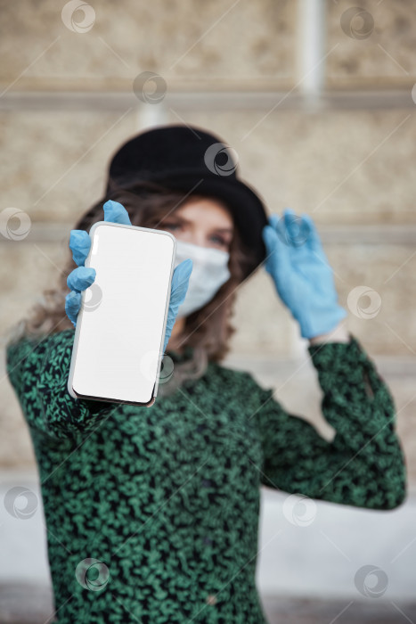 Скачать Симпатичная молодая женщина во французском стиле в медицинской маске и перчатках показывает мобильный телефон фотосток Ozero