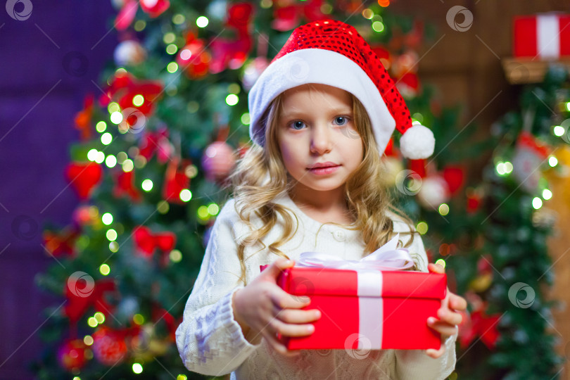 Скачать Маленькая девочка сидит у елки, держа в руках рождественский подарок В шляпе Санта-Клауса фотосток Ozero