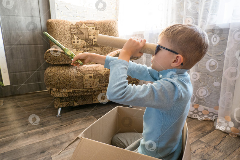 Скачать Игривый жизнерадостный ребенок, мальчик, заглядывающий в картонную трубочку, сидящий в коробке фотосток Ozero