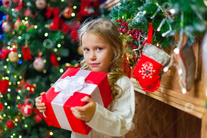 Скачать Маленькая девочка сидит у елки с рождественским подарком в руках фотосток Ozero