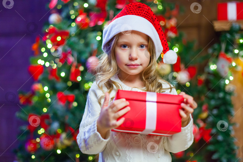 Скачать Маленькая девочка сидит у елки, держа в руках рождественский подарок В шляпе Санта-Клауса фотосток Ozero