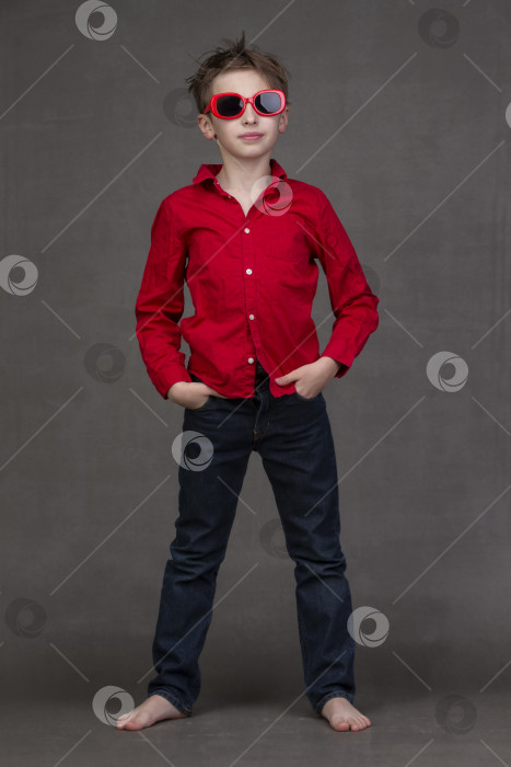 Скачать Вертикальный портрет стильного маленького мальчика в полный рост в красных солнцезащитных очках на сером фоне. фотосток Ozero