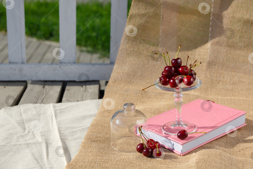 Скачать Сервировочная тарелка на ножках и мини-купол с ягодами вишни находятся на книге и на мешковине на деревянном полу фотосток Ozero