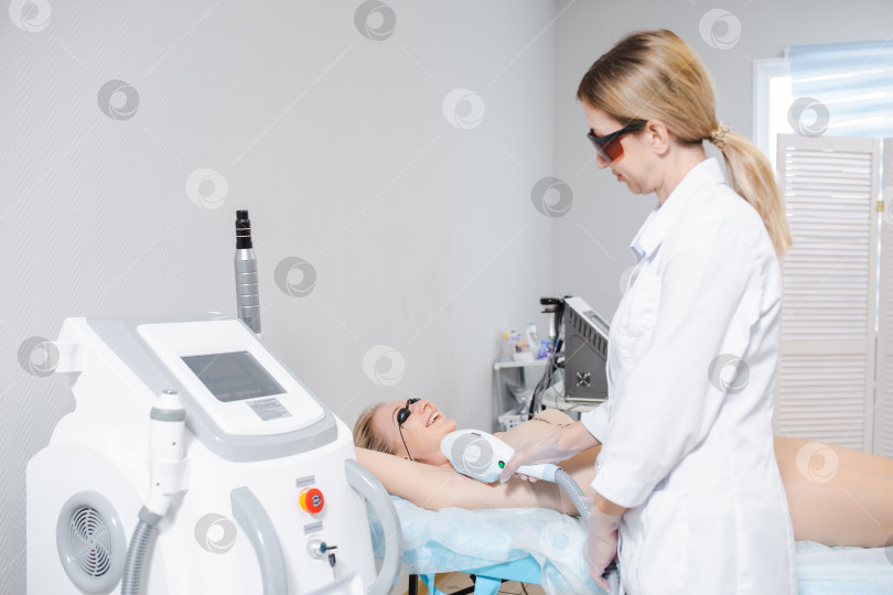 Скачать Фотография современного косметологического аппарата и женщины-дерматолога в защитных очках, выполняющей процедуру лазерной эпиляции подмышек молодой девушки фотосток Ozero
