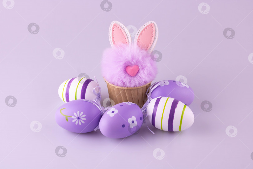 Скачать Сиреневый пушистый игрушечный кролик в форме для выпечки кексов, рядом с ним лежат пасхальные яйца. фотосток Ozero