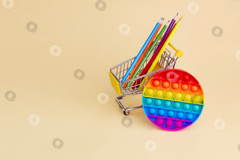 Скачать Популярная игра Pop it и цветные карандаши в тележке супермаркета на желтом фоне. фотосток Ozero