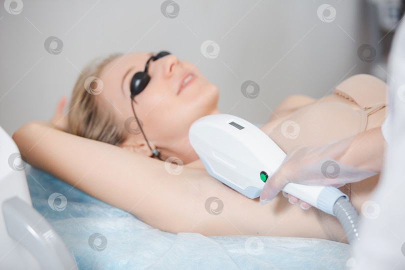 Скачать Фотография современного косметологического аппарата и женщины-дерматолога в защитных очках, выполняющей процедуру лазерной эпиляции подмышек молодой девушки фотосток Ozero
