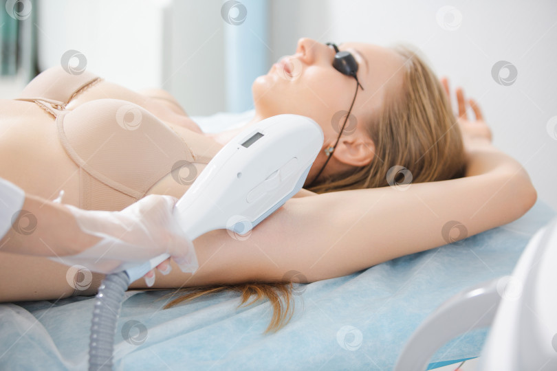 Скачать Крупный план устройства для лазерной эпиляции во время эпиляции подмышек молодой женщины, лежащей в защитных очках в косметологическом кабинете фотосток Ozero