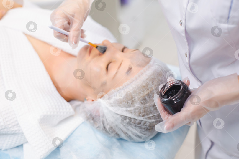 Скачать Руки косметолога, наносящего углеродный наногель на лицо молодой пациентки с помощью кисти, готовящейся к процедурам карбонового пилинга фотосток Ozero