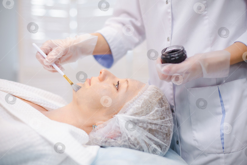 Скачать Вид сбоку на руки косметолога, наносящего углеродный наногель на лицо молодой пациентки с помощью кисти, готовящейся к процедурам углеродного пилинга фотосток Ozero