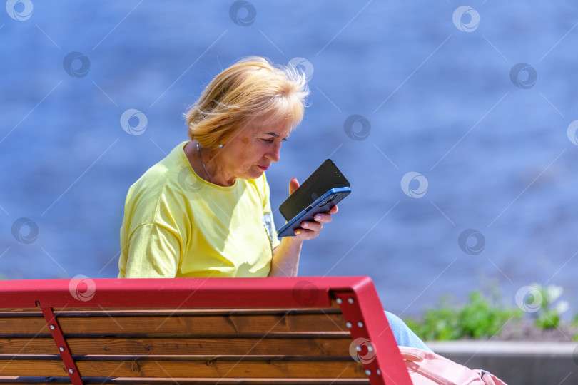 Скачать Череповец, Россия, 21 июля 2023 года Женщина в желтой футболке сидит на красной деревянной скамейке на фоне моря или реки, глядя в телефон. Интернет-общение и социальные сети. фотосток Ozero