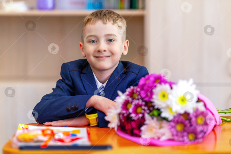 Скачать Портрет умного улыбающегося мальчика - ученика начальных классов в школьной форме, сидящего за школьной партой с букетом цветов. Праздник "День знаний - 1 сентября" фотосток Ozero