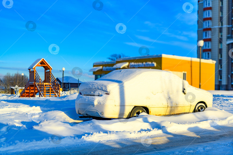 Скачать Белый автомобиль, покрытый снегом после метели и снегопада, припаркован на обочине дороги на городской улице. Сугробы и замерзшие автомобили холодной зимой.Яркий солнечный день. фотосток Ozero