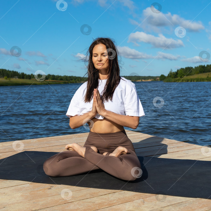 Скачать Привлекательная женщина, практикующая йогу, медитирующая, сидя в позе лотоса с намасте, тренирующаяся в спортивной одежде, сидя на коврике у озера солнечным утром фотосток Ozero