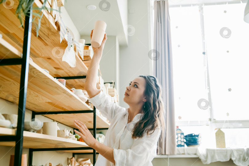 Скачать Молодая женщина-менеджер гончарной мастерской расставляет изделия на полке фотосток Ozero