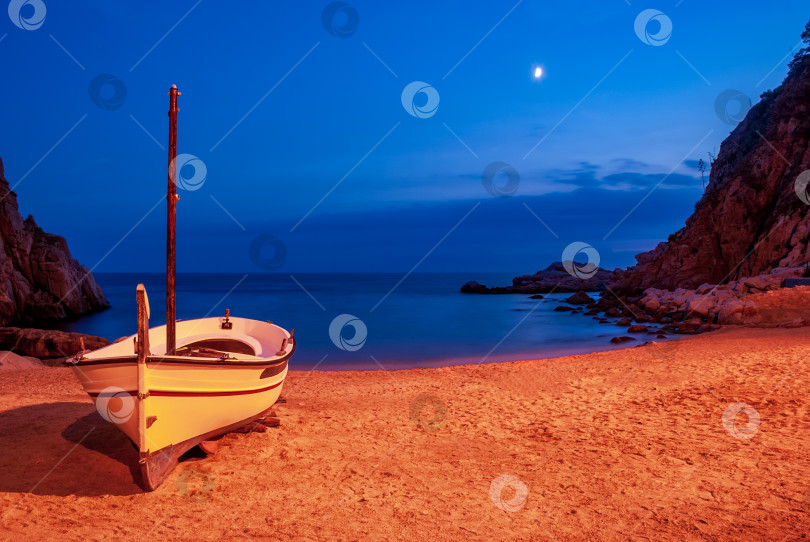 Скачать Лодка на песчаном пляже в заливе Тосса-де-Мар лунной ночью на фоне гор и голубого неба. фотосток Ozero
