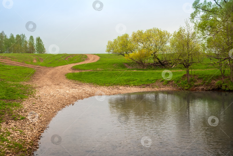 Скачать Сельская грунтовая дорога в сельской местности. Зеленые поля, деревья и небольшая река. Красивый весенний пейзаж. фотосток Ozero