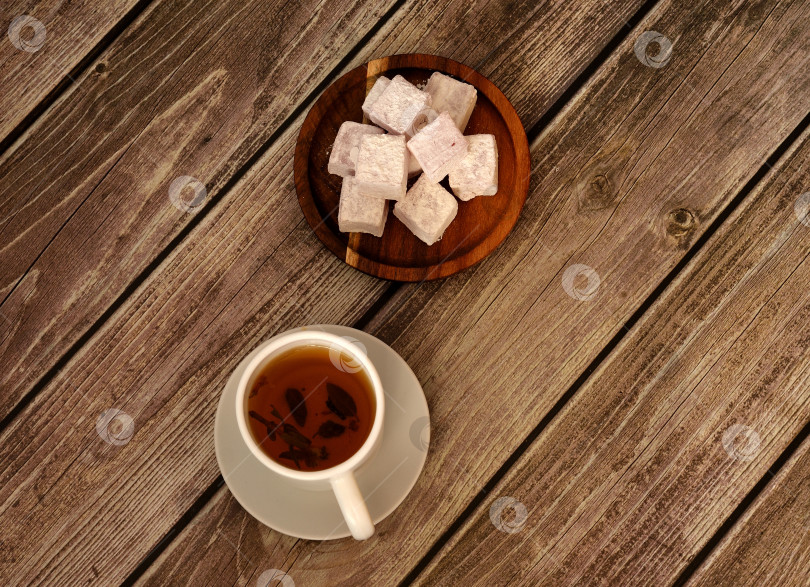 Скачать Белая фарфоровая чашка горячего чая на блюдце и круглая деревянная тарелка с кучей восточных сладостей в сахарной пудре на деревянном столе. фотосток Ozero