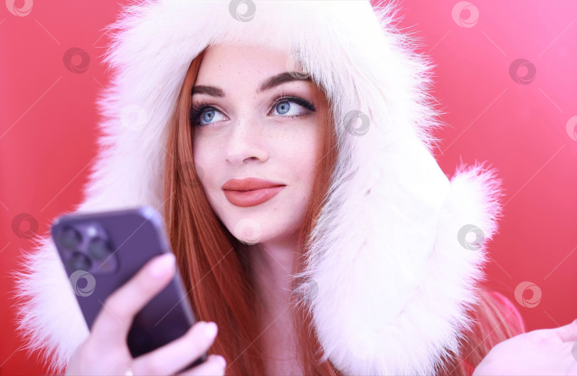 Скачать красивая девушка с рыжими волосами в белой пушистой шапочке на красном фоне делает селфи на мобильный телефон фотосток Ozero