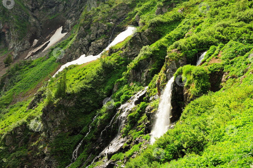 Скачать Небольшой водопад бурным потоком стекает с вершины горы, поросшей травой и кустарником, с остатками снега на склонах. фотосток Ozero