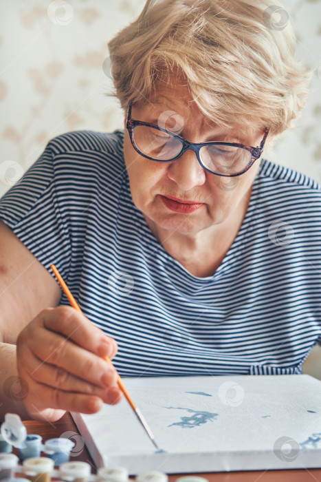 Скачать Привлекательная пожилая женщина рисует дома по номерам. Вдохновляющее хобби и арт-терапия. Избирательный подход фотосток Ozero