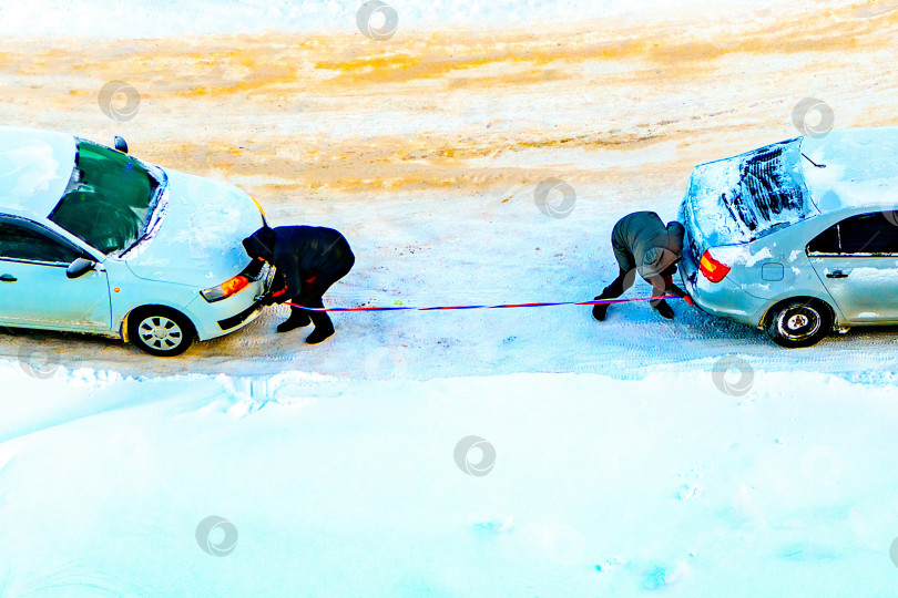 Скачать Водители прикрепляют буксирный трос к своим автомобилям, чтобы буксировать неисправный замерзший автомобиль морозной зимой.Вид сверху.Техническая помощь, буксировка неисправного автомобиля.Буксировка. фотосток Ozero