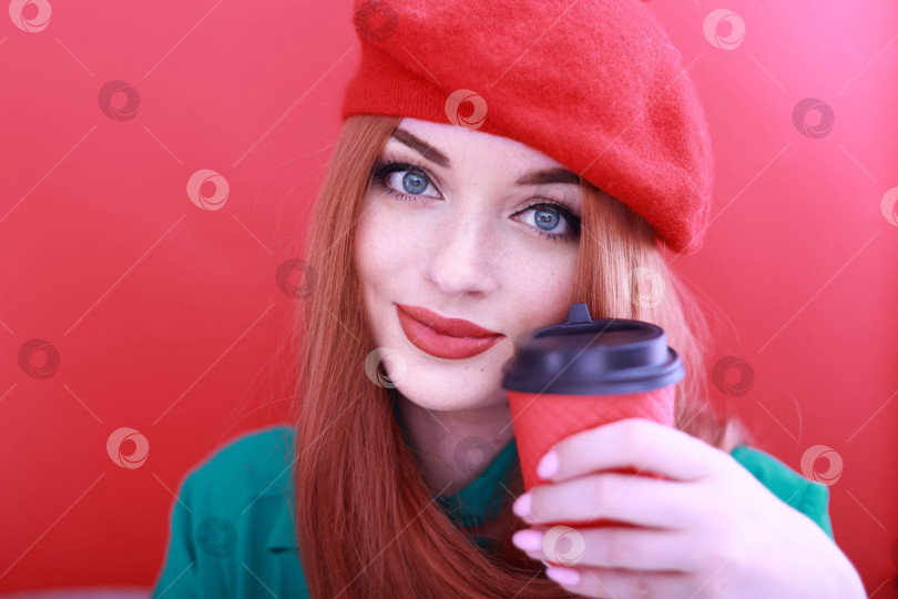 Скачать Романтичная молодая женщина, держащая бумажный красный стаканчик с кофе или чаем. рыжеволосая девушка в ярко-красном головном уборе. Выделено на красном фоне фотосток Ozero