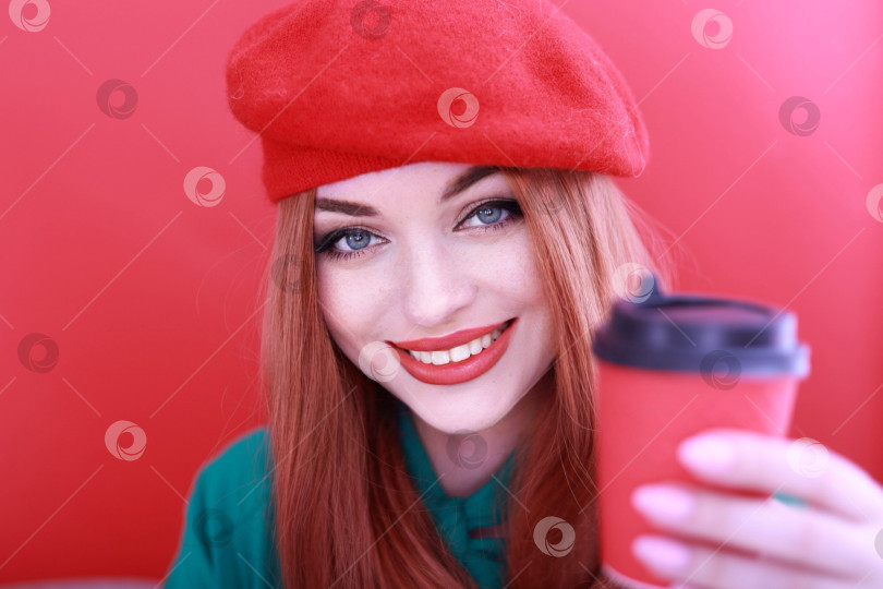 Скачать Романтичная молодая женщина, держащая бумажный красный стаканчик с кофе или чаем. рыжеволосая девушка в ярко-красном головном уборе. Выделено на красном фоне фотосток Ozero