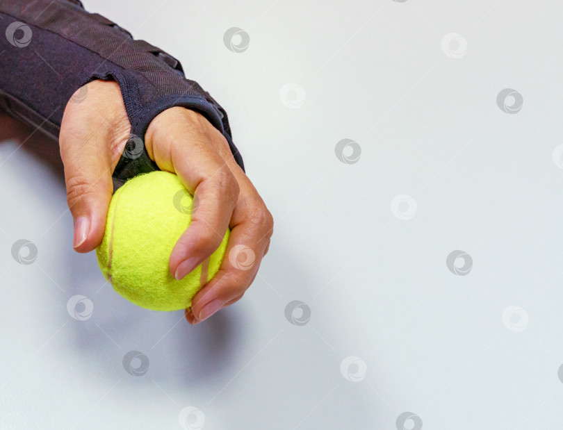 Скачать Сломанная рука в черном ортезе, держащая желтый теннисный мяч для тренировок и восстановления на светло-сером фоне фотосток Ozero