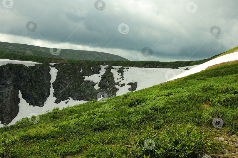 Скачать Пологий склон холма и отвесные скалы с остатками снега в пасмурный летний день. фотосток Ozero