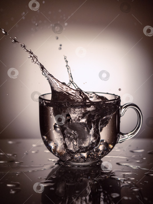 Скачать Лед был брошен в чашку с водой или прозрачной жидкостью. Брызги от напитка разлетелись из чашки в разные стороны и попали на отражающую поверхность стола. фотосток Ozero