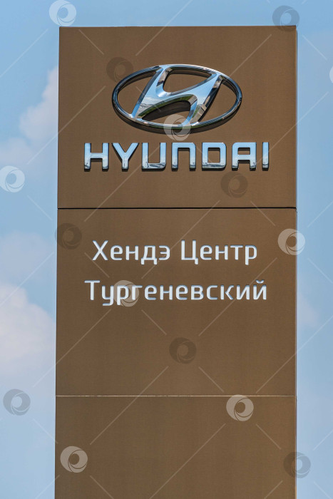 Скачать Логотип автопроизводителя Hyundai на коричневом вертикальном пилоне. Пилон с логотипом между шоссе и автостоянкой перед сервисным центром Hyundai в Адыгее. фотосток Ozero