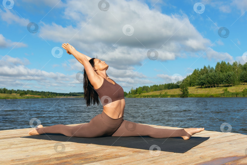 Скачать Стройная женщина, ведущая здоровый образ жизни, выполняет упражнение Хануманасана, продольный шпагат, тренируется в леггинсах и футболке, сидя на берегу озера теплым солнечным утром фотосток Ozero
