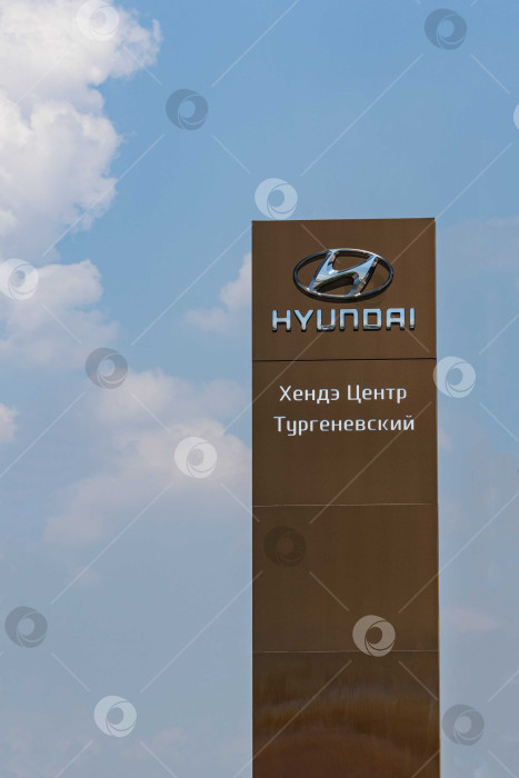 Скачать Логотип автопроизводителя Hyundai на коричневом вертикальном пилоне. Пилон с логотипом между шоссе и автостоянкой перед сервисным центром Hyundai в Адыгее. фотосток Ozero