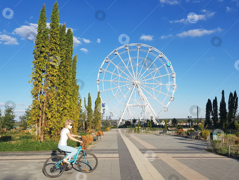 Скачать Девушка на велосипеде в черных очках и защитной маске едет по парку на фоне аттракциона Колесо обозрения фотосток Ozero