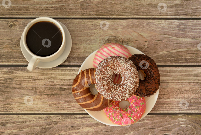 Скачать Чашка горячего черного кофе на блюдце и тарелка с горкой свежих пончиков в разноцветной глазури на деревянном столе. фотосток Ozero