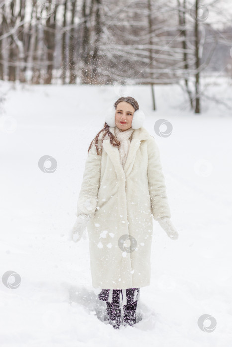 Скачать Счастливая молодая женщина в белой шубе, варежках и меховых наушниках на зимней прогулке в парке фотосток Ozero