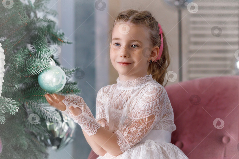 Скачать Симпатичная маленькая девочка-карапуз в белом праздничном платье сидит возле праздничной новогодней елки. Рождество фотосток Ozero