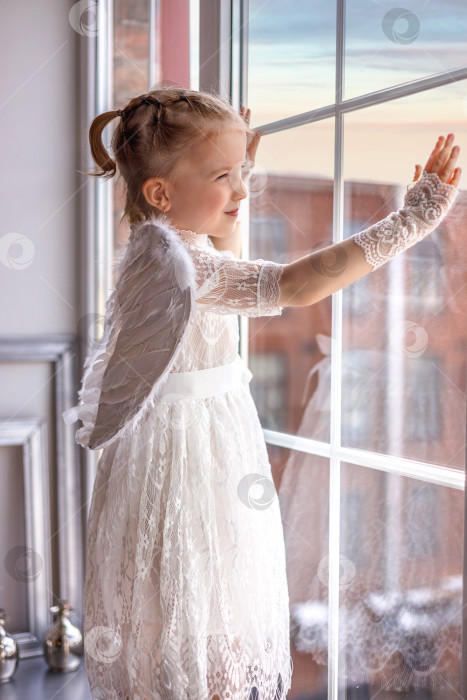 Скачать Маленький ангел стоит на подоконнике и смотрит в окно. Милая маленькая девочка-ангел смотрит и улыбается. фотосток Ozero