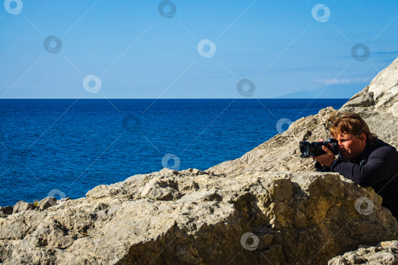 Скачать Молодой человек стоит на скалистом берегу и фотографирует мыс Алчак. Дикая природа возле древнего города, построенного генуэзцами. Огромные камни и валуны на фоне моря. фотосток Ozero